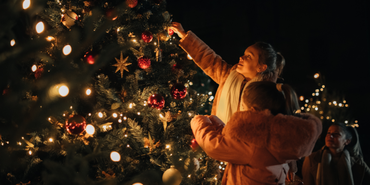 It's dark out with only the Christmas tree lights illuminating the people as they reach up to put ornaments on it.